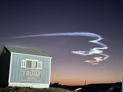 A small building with a sign supporting Trump 2020 sits in the foreground. Above it, a bright, white contrail or cloud formation curves dynamically against the backdrop of a dusky sky with a gradient from deep blue to orange near the horizon.