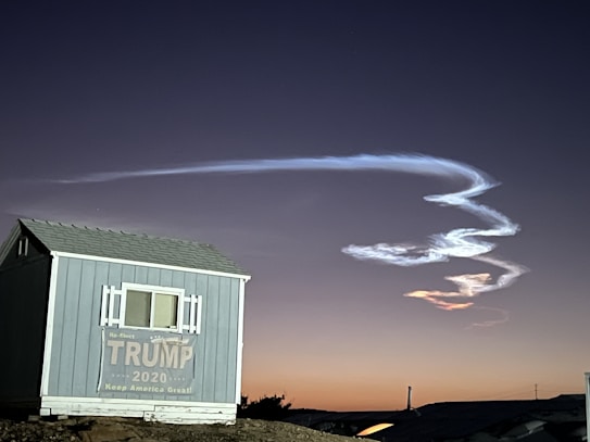 A small building with a sign supporting Trump 2020 sits in the foreground. Above it, a bright, white contrail or cloud formation curves dynamically against the backdrop of a dusky sky with a gradient from deep blue to orange near the horizon.
