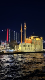 An illuminated mosque with two minarets is seen on the waterfront at night. In the background, a suspension bridge with red and white lighting stretches across the sky. The scene reflects in the slightly rippling water in the foreground.
