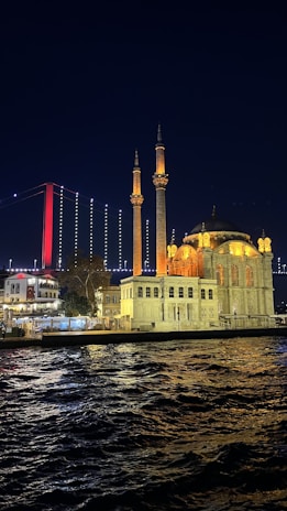 An illuminated mosque with two minarets is seen on the waterfront at night. In the background, a suspension bridge with red and white lighting stretches across the sky. The scene reflects in the slightly rippling water in the foreground.