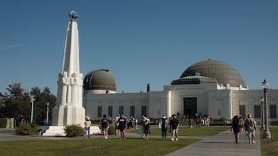 A large white building with a dome structure, identified as an observatory, stands against a clear blue sky. In front of the building, a tall white monument with figures on it is surrounded by people walking and enjoying the sunny day. The area is landscaped with grass and shrubs, and there's a sense of activity as visitors explore the site.