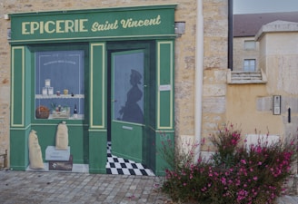 A painted mural on a stone wall depicts a storefront of a shop named 'EPICERIE Saint Vincent'. The mural includes a window displaying jars, baskets of vegetables, wine bottles, and other goods. A silhouette of a person is visible through the painted door. The mural is detailed, with a green and yellow color scheme, and is accompanied by pink flowers growing in front.