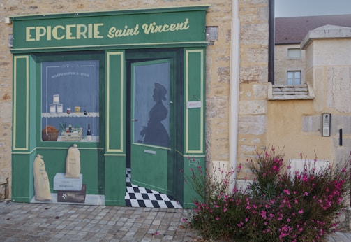 A painted mural on a stone wall depicts a storefront of a shop named 'EPICERIE Saint Vincent'. The mural includes a window displaying jars, baskets of vegetables, wine bottles, and other goods. A silhouette of a person is visible through the painted door. The mural is detailed, with a green and yellow color scheme, and is accompanied by pink flowers growing in front.