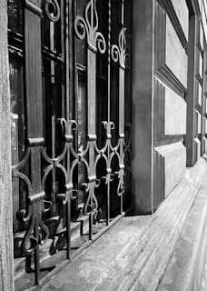 An ornate wrought iron fence with intricate curved patterns lines the exterior of a building. The wall of the building is composed of large stone blocks, adding to the classical architectural style. Light creates shadows on the wall, highlighting the details of the ironwork.