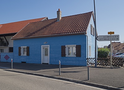 A small blue wooden house with brown shutters and a red-tiled roof is situated by the roadside. There is a directional sign indicating locations to Hombourg and Ottmarsheim. The house neighbors another building with a similar roof style. The sky is clear and there are some shrubs near the house.