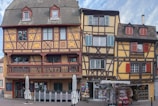Three traditional half-timbered buildings with intricate patterns and a contrasting color palette create a charming and historical appearance. The ground floors house a restaurant and a souvenir shop, and there are stacked chairs and closed umbrellas suggesting an outdoor seating area.