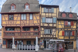 Three traditional half-timbered buildings with intricate patterns and a contrasting color palette create a charming and historical appearance. The ground floors house a restaurant and a souvenir shop, and there are stacked chairs and closed umbrellas suggesting an outdoor seating area.