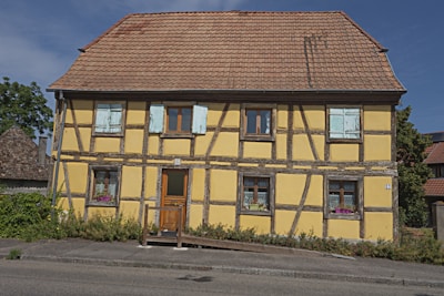 A traditional half-timbered house with a yellow exterior and a tiled roof. The building features wooden beams arranged in a grid pattern. Several windows have shutters, some painted in a faded light blue. Flower boxes with pink and purple flowers are seen on the window sills.