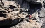 A person wearing a helmet and climbing gear is ascending a rugged, rocky cliff face. The sunlight casts shadows on the uneven surface of the rocks, highlighting their rough texture. The scene conveys a sense of adventure and challenge.