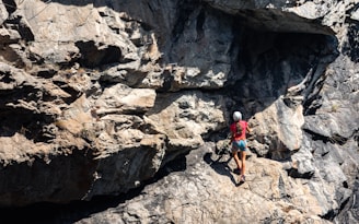 A person wearing a helmet and climbing gear is ascending a rugged, rocky cliff face. The sunlight casts shadows on the uneven surface of the rocks, highlighting their rough texture. The scene conveys a sense of adventure and challenge.
