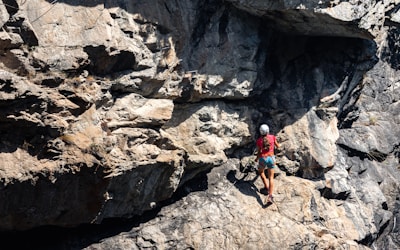 A person wearing a helmet and climbing gear is ascending a rugged, rocky cliff face. The sunlight casts shadows on the uneven surface of the rocks, highlighting their rough texture. The scene conveys a sense of adventure and challenge.