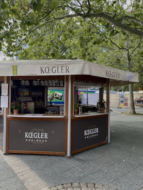 A kiosk with a canopy displaying the name 'Koegler' is situated in an outdoor setting. The kiosk is surrounded by large trees with green leaves, creating a serene atmosphere. Several potted plants decorate the counter, and wine glasses are visible hanging inside the kiosk.