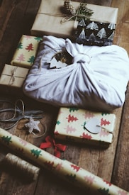 A collection of wrapped gifts is arranged on a wooden surface. Several presents are decorated with Christmas tree-patterned wrapping paper, and one prominent gift is wrapped in white cloth using the furoshiki technique. Various elements such as a spool of twine, a pair of scissors, a Christmas tree ornament, and a roll of wrapping paper with a festive design are also visible.