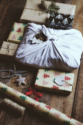 A collection of wrapped gifts is arranged on a wooden surface. Several presents are decorated with Christmas tree-patterned wrapping paper, and one prominent gift is wrapped in white cloth using the furoshiki technique. Various elements such as a spool of twine, a pair of scissors, a Christmas tree ornament, and a roll of wrapping paper with a festive design are also visible.
