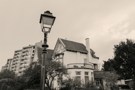 A black and white photograph features a large house with Tudor-style architecture, surrounded by trees. In the foreground, there's a vintage street lamp. In the background, an apartment building can be seen, providing contrast between modern and classic architecture. The sky is overcast, adding a moody atmosphere.