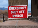 A red emergency shut-off switch sign is mounted on a wooden post. The sign is vibrant with bold, white text. The background features a blurred landscape with hints of mountains and sky.