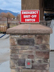 A stone pillar outdoors is topped with a sign that reads 'Emergency Shut-Off Switch' in bold white letters on a red background. Below the sign, there is a covered button with a red circle, presumably the emergency switch. In the background, there are trees, part of a building, and distant mountains under a cloudy sky.