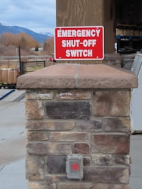 A stone pillar outdoors is topped with a sign that reads 'Emergency Shut-Off Switch' in bold white letters on a red background. Below the sign, there is a covered button with a red circle, presumably the emergency switch. In the background, there are trees, part of a building, and distant mountains under a cloudy sky.