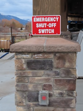 A stone pillar outdoors is topped with a sign that reads 'Emergency Shut-Off Switch' in bold white letters on a red background. Below the sign, there is a covered button with a red circle, presumably the emergency switch. In the background, there are trees, part of a building, and distant mountains under a cloudy sky.