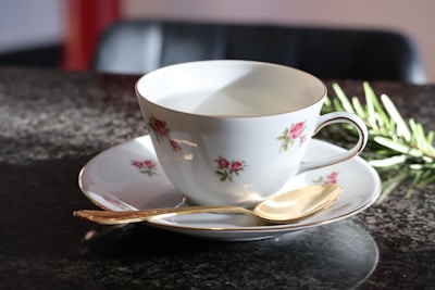 A porcelain teacup and saucer adorned with delicate pink floral patterns sit on a granite countertop. A gold spoon is placed neatly on the saucer. The scene is softly lit, providing a warm ambiance. In the background, a sprig of rosemary adds a touch of greenery.