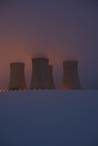 Four cooling towers rise against a dusk or dawn sky, with faint smoke or steam visible at the top. The scene is enveloped in a mix of dark blue and an orange glow from either the setting or rising sun. Power lines stretch across the image, and the ground is covered in snow, creating a stark contrast with the structures.