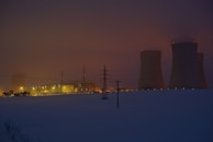 A power plant with cooling towers and smokestacks is illuminated at dusk. The surrounding area appears to be covered in snow, and the sky is dark with a reddish hue, suggesting evening lighting. Power lines are visible in the foreground.