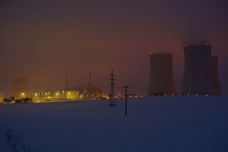 A power plant with cooling towers and smokestacks is illuminated at dusk. The surrounding area appears to be covered in snow, and the sky is dark with a reddish hue, suggesting evening lighting. Power lines are visible in the foreground.