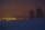 A power plant with cooling towers and smokestacks is illuminated at dusk. The surrounding area appears to be covered in snow, and the sky is dark with a reddish hue, suggesting evening lighting. Power lines are visible in the foreground.