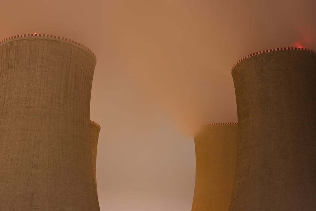 Hyperbolic natural-draft evaporative cooling towers at a thermal power station at night, illuminated against an atmospheric foggy sky — visualising the industrial scale of heat rejection these systems handle. Photo: Lukáš Lehotský / Unsplash