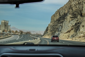 A road with several vehicles traveling on a highway with multiple lanes. On the right is a rocky hillside, and on the left, there are industrial structures, possibly factories or silos. The landscape in the background includes distant hills under a clear blue sky. The image is taken from inside a vehicle, evidenced by the dashboard and car interior framing the bottom part of the picture.