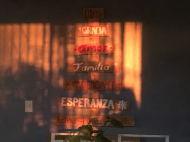 Wooden plaques are mounted on a wall, displaying words like 'Dios', 'Gracia', 'Amor', 'Familia', and more in Spanish. Sunlight casts warm shadows across the wall, enhancing the cozy atmosphere.
