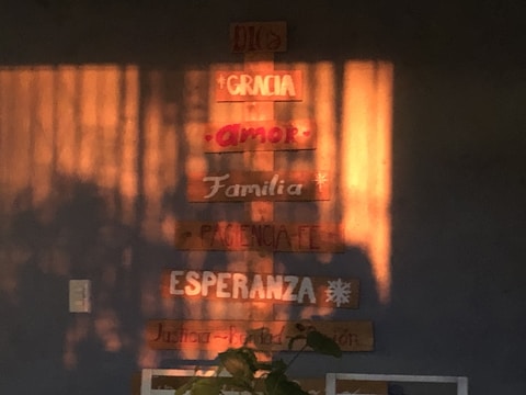 Wooden plaques are mounted on a wall, displaying words like 'Dios', 'Gracia', 'Amor', 'Familia', and more in Spanish. Sunlight casts warm shadows across the wall, enhancing the cozy atmosphere.