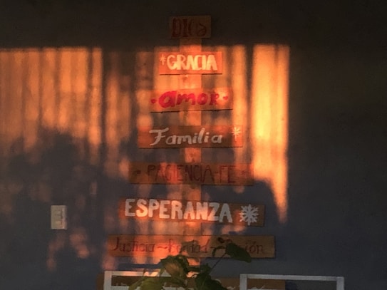 Wooden plaques are mounted on a wall, displaying words like 'Dios', 'Gracia', 'Amor', 'Familia', and more in Spanish. Sunlight casts warm shadows across the wall, enhancing the cozy atmosphere.
