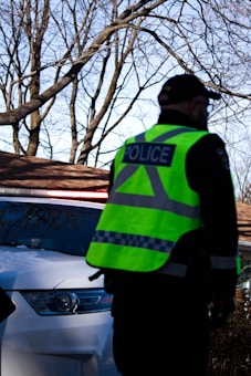 A police officer wearing a bright reflective vest stands in front of a white police car with flashing lights. Bare tree branches and part of a house roof are visible in the background.