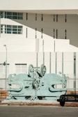 A large, industrial machine with gears and mechanical parts is positioned outdoors in front of a modern building. The building features geometric architectural elements with multiple small windows and a smooth white facade. A sparse urban setting with minimal vegetation surrounds the scene.