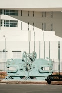 A large, industrial machine with gears and mechanical parts is positioned outdoors in front of a modern building. The building features geometric architectural elements with multiple small windows and a smooth white facade. A sparse urban setting with minimal vegetation surrounds the scene.
