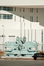 A large, industrial machine with gears and mechanical parts is positioned outdoors in front of a modern building. The building features geometric architectural elements with multiple small windows and a smooth white facade. A sparse urban setting with minimal vegetation surrounds the scene.