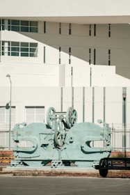A large, industrial machine with gears and mechanical parts is positioned outdoors in front of a modern building. The building features geometric architectural elements with multiple small windows and a smooth white facade. A sparse urban setting with minimal vegetation surrounds the scene.