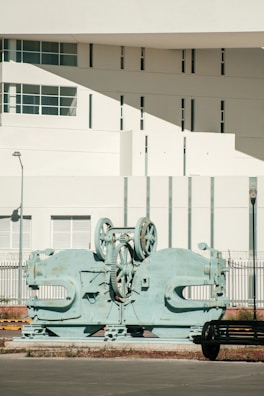 A large, industrial machine with gears and mechanical parts is positioned outdoors in front of a modern building. The building features geometric architectural elements with multiple small windows and a smooth white facade. A sparse urban setting with minimal vegetation surrounds the scene.