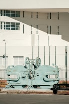 A large, industrial machine with gears and mechanical parts is positioned outdoors in front of a modern building. The building features geometric architectural elements with multiple small windows and a smooth white facade. A sparse urban setting with minimal vegetation surrounds the scene.