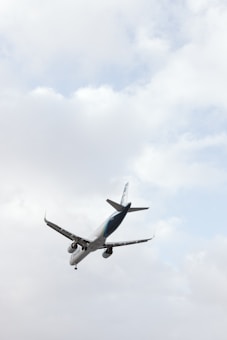 An aircraft is captured in mid-air against a backdrop of a cloudy sky. The angle of the photo emphasizes the airplane's underside, with the wings and engines clearly visible.