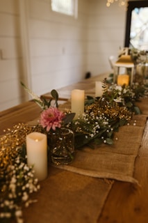 A wooden table is elegantly decorated with a rustic burlap runner, floral arrangements of white and pink flowers, and lush greenery. Candles are placed in between the flowers, emitting a soft, warm glow. In the background, fairy lights add a twinkling effect, adding to the cozy ambiance. A glass vase and a lantern are part of the decoration, enhancing the charming, rustic feel.