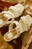 A pair of elegant sandals with braided straps adorned with pearls, resting on a wooden surface. Soft fabric pieces in warm tones surround the shoes, adding a touch of luxury and style.