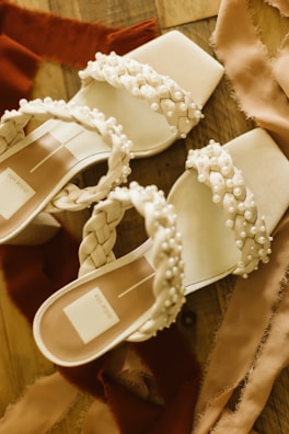 A pair of elegant sandals with braided straps adorned with pearls, resting on a wooden surface. Soft fabric pieces in warm tones surround the shoes, adding a touch of luxury and style.