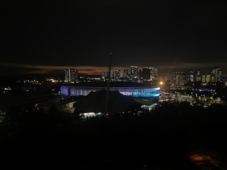 A nighttime cityscape featuring a brightly lit stadium surrounded by high-rise buildings. The stadium emits vibrant colors, and there are scattered lights from buildings and streets. The skyline is silhouetted against a dark sky with a faint glow near the horizon.