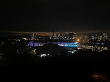A nighttime cityscape featuring a brightly lit stadium surrounded by high-rise buildings. The stadium emits vibrant colors, and there are scattered lights from buildings and streets. The skyline is silhouetted against a dark sky with a faint glow near the horizon.
