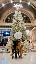 A tall, elaborately decorated Christmas tree stands in the center of a spacious, elegantly designed interior with a high arched ceiling. The tree is adorned with gold and white decorations, including large stars, ribbons, and floral arrangements. In front of the tree, there are three people posing for a photo: an adult man, a child, and an adult woman dressed in casual attire. They are all smiling and standing on a polished marble floor. A red rope barrier partially encircles the tree. Signage for 'Nustar Resort Cebu' is prominently displayed on the tree.