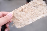 A close-up of a hand holding a textured piece of handmade paper with visible fibers and specks. The paper appears fibrous and natural, with a light beige color contrasting with the soft focus background.