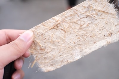 A close-up of a hand holding a textured piece of handmade paper with visible fibers and specks. The paper appears fibrous and natural, with a light beige color contrasting with the soft focus background.
