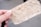 A close-up of a hand holding a textured piece of handmade paper with visible fibers and specks. The paper appears fibrous and natural, with a light beige color contrasting with the soft focus background.